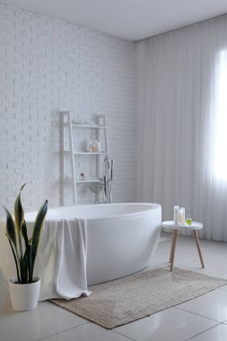 Interior of light bathroom with bathtub, table and shelving unit