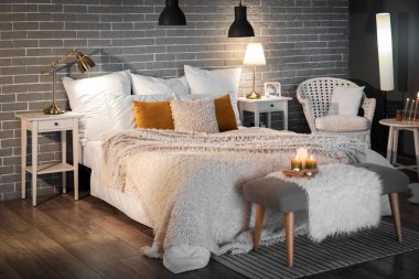 Interior of bedroom with cozy blankets on bed, burning candles and glowing lamps late in evening