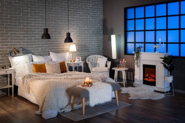 Interior of bedroom with cozy blankets on bed, burning candles and glowing lamps late in evening
