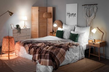 Interior of bedroom with brown checkered blankets on bed and glowing lamps late in evening
