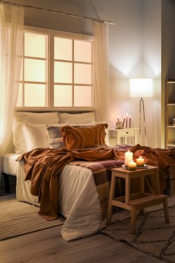Interior of bedroom with brown checkered blankets on bed, burning candles and glowing lamp late in evening