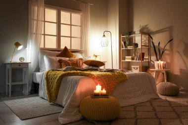 Interior of bedroom with cozy blankets on bed, burning candles and glowing lamp late in evening