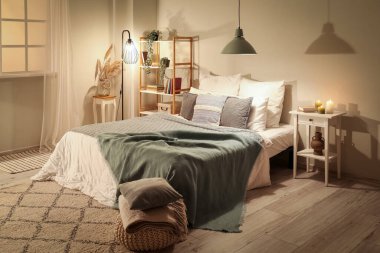 Interior of bedroom with green blankets on bed, burning candles and glowing lamps late in evening