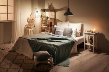 Interior of bedroom with green blankets on bed, burning candles and glowing lamps late in evening