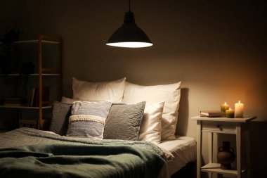 Interior of bedroom with green blankets on bed, burning candles and glowing lamp at night