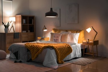 Interior of bedroom with knitted plaid on bed and glowing lamps at night