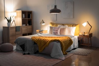 Interior of bedroom with knitted plaid on bed and glowing lamps late in evening