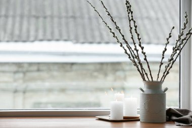 Vase with willow branches and burning candles on windowsill in room