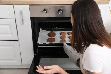 Young woman taking baking tray with cookies from electric oven in kitchen, closeup