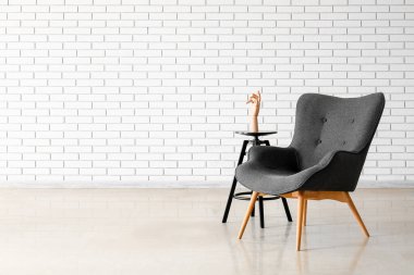 Stylish grey armchair and end table with wooden hand near white brick wall