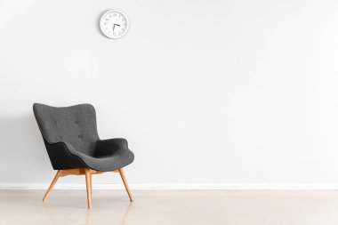 Stylish grey armchair and clock near white wall