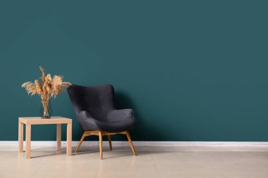 Stylish grey armchair and vase with pampas grass on table near green wall