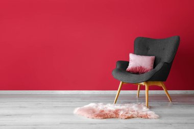 Stylish grey armchair, cushion and pink fur rug near red wall