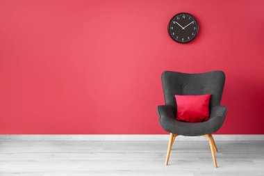 Stylish grey armchair, cushion and clock near red wall