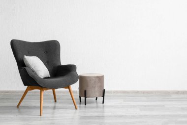 Stylish grey armchair, cushion and pouf near white wall
