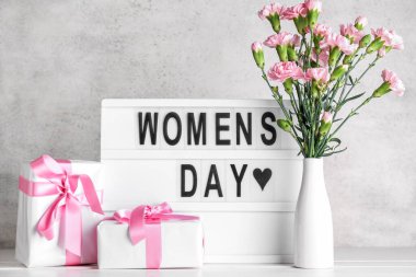 Text WOMEN'S DAY, gift boxes and vase with beautiful carnation flowers on table near grey wall