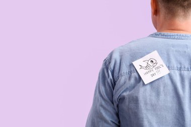 Sticky paper with text HAPPY FOOL'S DAY on back of mature man against lilac background, closeup