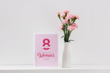 Vase with carnation flowers and greeting card for Women's Day on shelf in light room