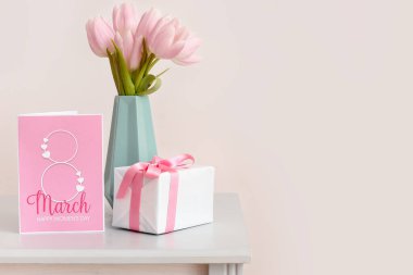 Vase with tulips, gift and greeting card for Women's Day on shelf near light wall