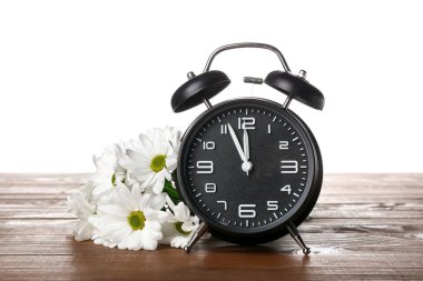 Beautiful chamomile flowers and alarm clock on wooden table against white background