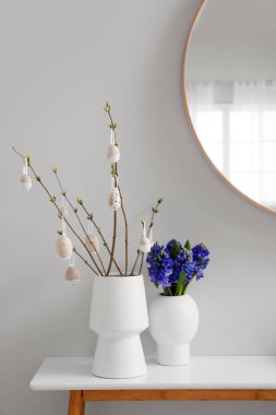 Vases with tree branches, Easter eggs and flowers on table in room