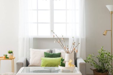Vase with tree branches, Easter eggs and rabbits on table in light living room