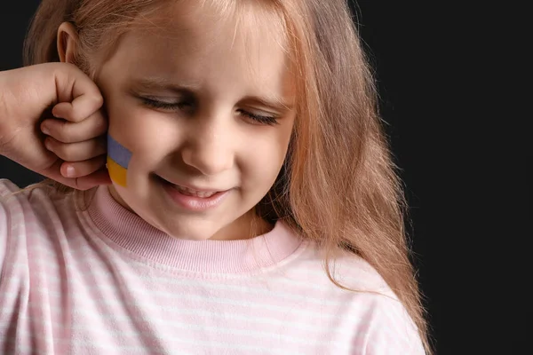 Little girl with drawn flag of Ukraine crying on black background, closeup