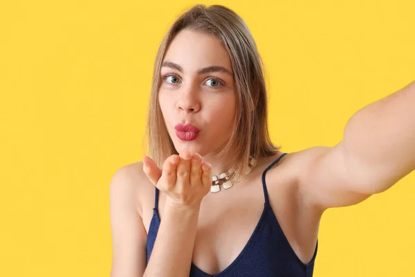 Young woman with necklace blowing kiss on yellow background, closeup