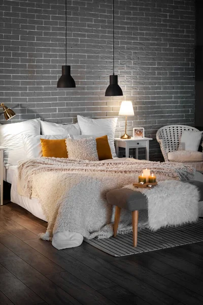Interior of bedroom with cozy blankets on bed, burning candles and glowing lamps late in evening