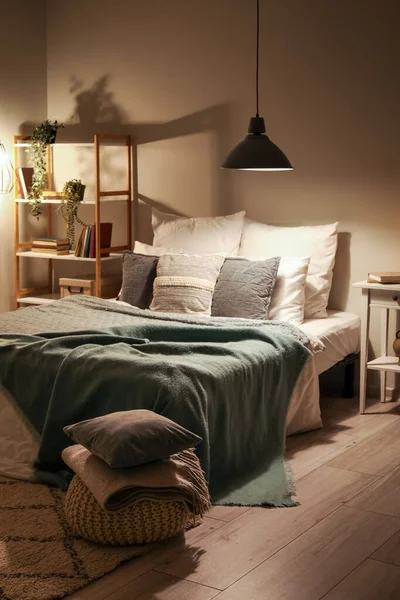 Interior of bedroom with cozy blankets on bed and glowing lamp late in evening