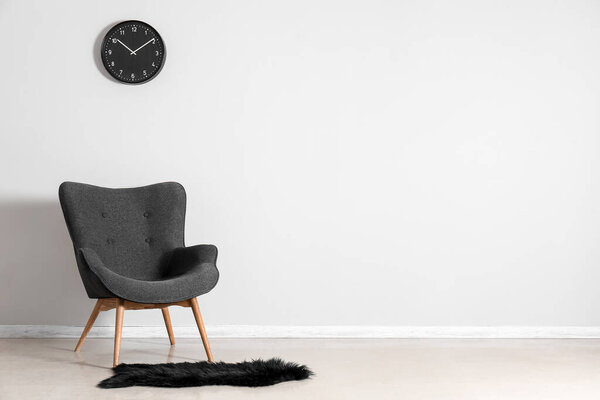 Стильное серый кресло, часы и черный ковер меха возле белой стены