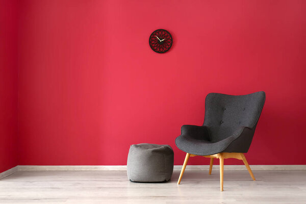 Стильное серое кресло, пуф и часы возле красной стены