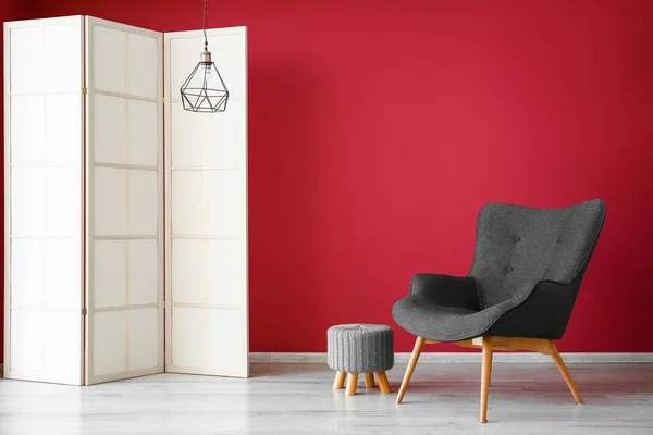 Stylish grey armchair, folding screen, pouf and lamp near red wall