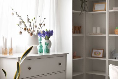 Vases with tree branches, Easter eggs, flowers and rabbits on chest of drawers in room