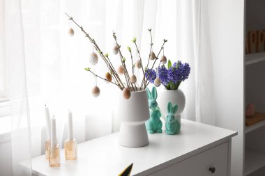Vases with tree branches, Easter eggs, flowers and rabbits on chest of drawers in room