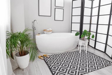 Interior of light bathroom with bathtub, blank frames and houseplants