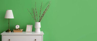 Chest of drawers with willow branches in vase, clock, books and lamp near green wall. Banner for design
