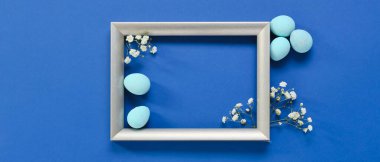 Beautiful Easter composition with blank frame, painted eggs and flowers on blue background