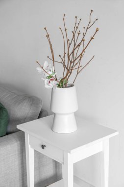 Vase with tree branches and flowers on table in light living room
