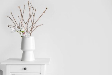 Vase with tree branches and flowers on table near light wall