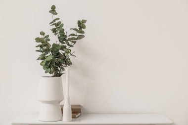 Vase with eucalyptus branches, burning candle and books on table near light wall