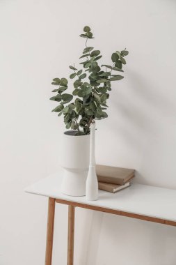 Vase with eucalyptus branches, burning candle and books on table near light wall