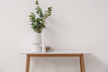 Vase with eucalyptus branches, burning candle and books on table near light wall