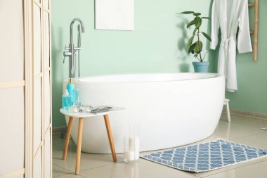 Interior of bathroom with modern bathtub and bath accessories near green wall
