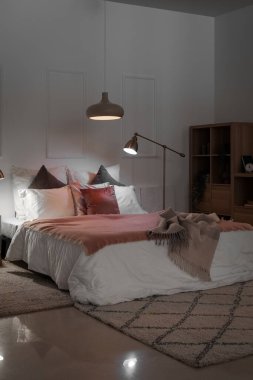 Interior of bedroom with blankets on bed and glowing lamps at night
