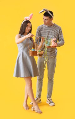 Happy young couple in bunny ears with Easter eggs and rabbits on yellow background