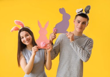 Happy young couple in bunny ears with paper Easter rabbits on yellow background