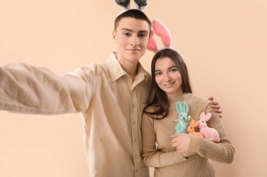 Happy young couple in bunny ears with Easter rabbits taking selfie on beige background