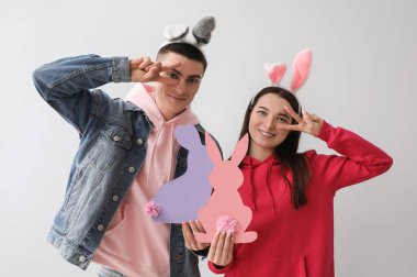 Young couple in bunny ears with paper Easter rabbits showing victory gesture on grey background