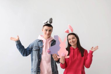 Young couple in bunny ears with paper Easter rabbits showing something on grey background